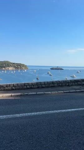 The video captures a scenic view of a bay dotted with numerous boats and yachts under a clear blue sky. In the foreground, a paved road with white markings runs parallel to a stone wall. Beyond the wall, the tranquil blue water of the bay stretches out, with a cluster of sailboats and smaller vessels anchored. Further in the distance, a large luxury yacht or cruise ship is visible, alongside more boats. The bay is bordered by lush green, hilly land on either side, with some buildings visible nestled within the landscape. The lighting suggests it is daytime, likely morning or afternoon, with the sun casting a subtle shadow on the road. There are no people visible in the frame.