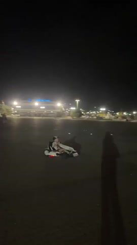 At night, a person in a kart is driving on a paved surface. The person is wearing a white jacket with black accents and dark pants. The kart has red lights on the back. In the background, there is a city skyline with lights.