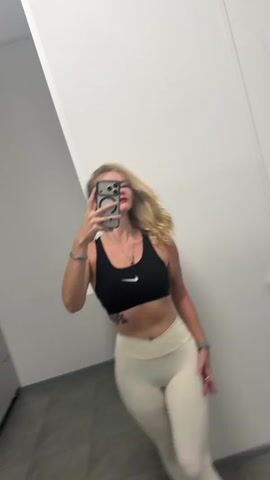 A blonde woman, wearing a black Nike sports bra and off-white leggings, poses for a mirror selfie. She holds her phone to take the picture, her face visible in the reflection. She has a tattoo on her left hip and is wearing jewelry on her left wrist and neck. The setting appears to be a gym or changing room, with tiled flooring and plain walls. The lighting suggests it could be daytime, but it's difficult to determine the exact time of day. The woman's expression is neutral, possibly indicating a focus on presenting herself in the outfit. There is no visible text in the media file.