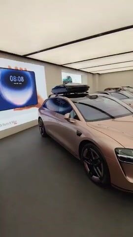 This is a brief, stationary video recording of the interior of a showroom, likely a car dealership. The focus is on a rose-gold colored electric car with a black roof rack and cargo box. The car is parked indoors with a dark floor and a white ceiling with strip lighting. To the left of the car, a large screen displays an advertisement, featuring a hand holding a blue smartphone and a tablet with a clock showing 08:08. Other cars are visible in the background, also parked indoors. The video provides a clear view of the car's front and side profile.