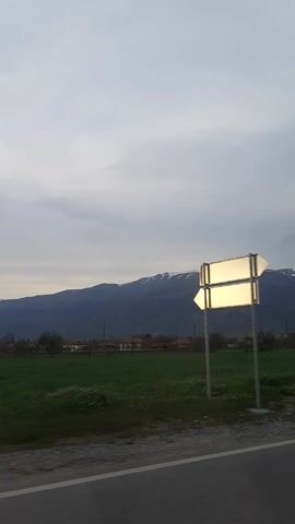 The video depicts a mountain range from a low angle, possibly taken from a moving vehicle. The sky is overcast with clouds. Green fields flank the mountain on the right side of the frame, with some buildings and trees visible in the distance. A road is on the rightmost part of the frame. The music appears to be playing loudly in the background, but the content is illegible.