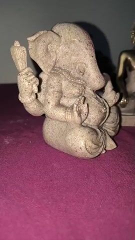 The video shows two statues positioned on a dark pink surface. In the foreground, a beige statue of Ganesha is visible from the side, holding an object in its upper right hand. To the right of Ganesha, a statue of Buddha is seen from the front. This Buddha statue is golden and beige and is seated in a meditative pose. Both statues appear to be made of a rough, granular material. Behind the Buddha statue, there are some decorative elements, including what looks like driftwood and a textured object resembling a pinecone or a cluster of scales. The background is a plain, light-colored wall with shadows cast by the statues and decorative elements.