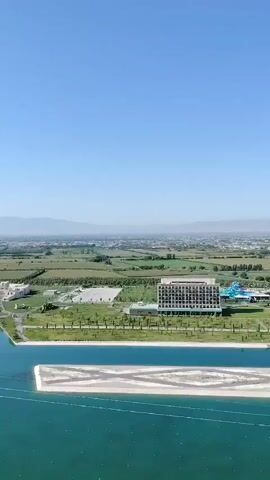 The video shows an aerial view of a resort area on a clear, sunny day. In the foreground, there is a large body of turquoise water with a sandy island in the middle. Beyond the water, there is a wide, blue artificial channel, likely a man-made lake or river, with a white sandy embankment on either side. Along the embankment of this channel, there are several small boats and what appear to be buoys or markers in the water. Further back, there are two modern hotel buildings, one with a darker facade and balconies, and another with a lighter, more ornate design. Between the hotels, there is a water park with blue slides. Beyond the hotels and water park, there is a green landscape with trees and fields. In the far distance, a city skyline with buildings and mountains can be seen under a clear blue sky. The camera appears to be moving and panning across the scene, offering a sweeping vista of the resort and its surroundings.