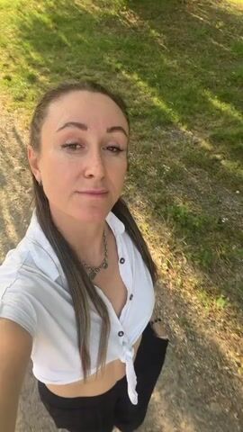 A woman with brown hair pulled into a ponytail, wearing a white tied-up shirt and black pants, walks while holding a phone and filming herself. She is outdoors on a path next to a grassy area and a body of water. The sun casts shadows on the ground.
