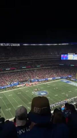 The video captures a moment from a football game at MetLife Stadium. The stadium is packed with spectators under the night sky. The camera angle is slightly elevated, looking down on the football field. A football game is in progress on the field. A man wearing a "Giants" hat stands in the foreground with others.
