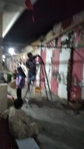 The video, filmed in Rambipuji, Indonesia, appears to be shot in the evening or at night due to the artificial lighting. The scene is an outdoor covered area, possibly a walkway or corridor, with a worn, peeling mural on the wall. The mural features red and white stripes and circular designs.

In the foreground, a person stands on an aluminum ladder, engaged in some activity near the ceiling, possibly painting or repairing. They are wearing a dark t-shirt with the number "33" in a white font and denim shorts. Below the ladder, another person is partially visible, wearing a plaid shirt and dark pants, and is looking upwards. Further in the background, other individuals and objects are dimly lit, including what appears to be a small yellow chair. The overall impression is one of an informal, possibly residential or commercial back area where maintenance or work is being carried out. The lighting is somewhat dim, creating shadows and highlighting the textures of the peeling paint on the wall. No specific weather conditions are discernible.