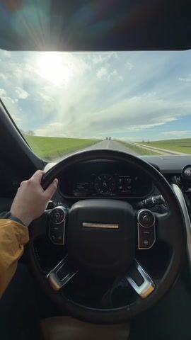 The video shows a driver's perspective from inside a Range Rover, speeding along a highway. The driver's left hand is visible on the steering wheel. The speedometer reads 78 mph. The landscape outside is green and mostly flat, with a bright, sunny sky with some clouds. There are a few vehicles visible in the distance on the highway. The sun is in front, creating a glare.