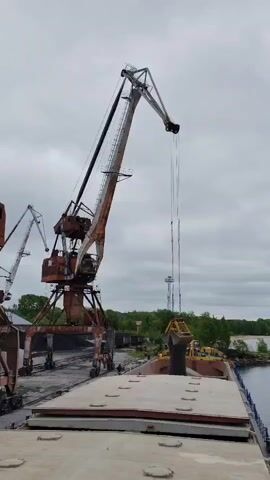 The video depicts an industrial port scene under an overcast sky, likely in Medvezhyegorsk, Russia. The primary activity involves a large, weathered gantry crane, numbered "42" on its base, actively unloading bulk material from the quay into the open hold of a large cargo vessel, possibly a barge or bulk carrier.

The crane's grab descends into the ship's hold, collects material (which appears to be dark, likely coal, gravel, or ore), lifts it, and then tips it out, filling the hold. Another similar, but smaller, gantry crane is partially visible to the left, also positioned alongside the quay, with piles of bulk material visible behind it.

The setting is characterized by heavy industrial equipment, a waterfront with what appears to be a river or lake, and a distant treeline of green foliage. The weather is cloudy and grey, suggesting a dull day. No specific time of day is discernible beyond daylight. There are no clearly identifiable people, though small figures might be present near the base of the cranes in the background. The overall impression is one of ongoing cargo operations at a working port.