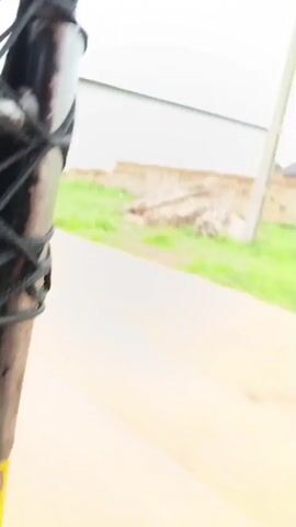 The video shows a sequence of shots taken from a moving vehicle, likely a car. The first few shots display an unfinished construction site with piles of bricks and bundles of what appear to be reeds or straw. There is a partially built structure with a white facade and a dark roof in the background. The ground is a mix of dirt and grass.

As the video progresses, the perspective shifts to show a more developed area. One shot captures a building with multiple floors and balconies, possibly a commercial or residential complex. Cars are parked in the foreground, and a motorbike is visible further back. Another shot focuses on a building with a sign that reads "BRIGHTSUN (IN) PLAZA". Electrical poles and wires are prominent in the scene.

The final shot shows a street with more developed houses and buildings on either side. Two people are walking away from the camera down the street. The environment appears to be somewhat rural or in a developing urban area, with dirt roads and partially constructed buildings interspersed with more finished structures. The sky is overcast.