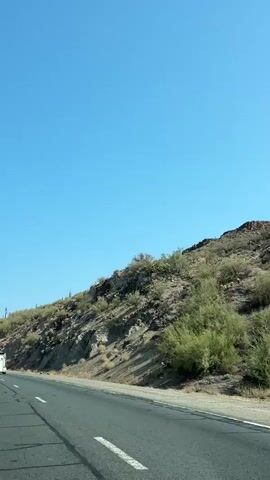 A clear blue sky stretches above a desert landscape. A paved road, with broken white lines marking lanes, curves to the left. On the right side of the road, a steep, rocky hillside is covered with sparse desert vegetation, including bushes and cacti. A white truck travels on the road, moving away from the camera. A silver car is visible in the lower right corner of the frame, also moving away. The sun casts shadows on the hillside, indicating it is daytime.