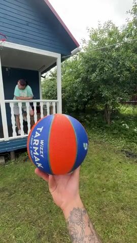 The video shows a person holding a basketball on their finger, spinning it. The ball is orange and blue with the text "TARMAK" and "BASKETBALL LIGHT & SOFT" visible. The person has a tattoo on their arm. In the background, a blue house with a white porch and railing is visible. A woman is standing on the porch, leaning on the railing and looking towards the person. The setting appears to be outdoors, with green grass and trees. The weather appears to be overcast. The time of day is not explicitly stated but the lighting suggests it could be daytime.
