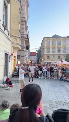 Two men are performing a street dance for a crowd gathered in a cobblestone plaza. The man in the foreground, wearing a light-colored shirt and shorts, is dancing in a crouched position with his arms extended. The man in the background, wearing a red bandana, white shirt, and shorts, stands with his arms at his sides. The crowd watches intently, some with hands raised. Buildings with light-colored facades line the plaza, and a clear blue sky is visible above. The time is early evening, indicated by the soft lighting. Several bags and other personal items are on the ground near the performers.
