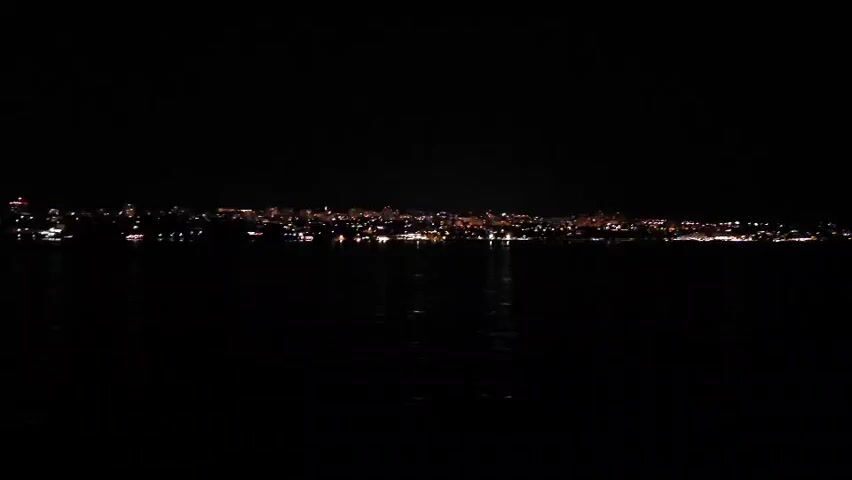 A wide shot captures a dark expanse of water in the foreground. Beyond the water, a long, low-lying cityscape is illuminated by a multitude of lights, suggesting a town or city at night. The lights stretch horizontally across the frame, with some appearing as individual points and others forming a more continuous glow. Reflections of these lights shimmer on the surface of the water, creating streaks of illumination that mirror the distant urban glow. The sky above is completely black, devoid of stars or moon, emphasizing the darkness of the environment and the isolation of the illuminated city. The overall scene is still and quiet, evoking a sense of nighttime tranquility or perhaps a remote, coastal settlement.