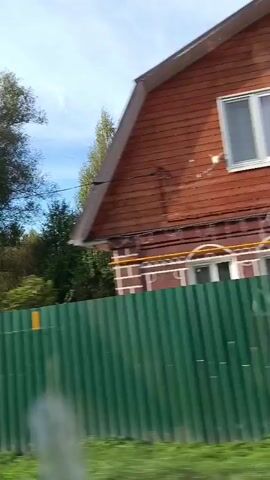 The video shows a view from a moving vehicle. The camera is positioned to capture the side of the road and the sky. In the foreground, there is a green corrugated fence. Behind the fence, trees with green and yellow foliage are visible. The sky is blue with scattered clouds. There are power lines and a utility pole visible in the distance. A car's side mirror is also visible, reflecting a person holding a phone, presumably taking the video. The road appears to be unpaved.