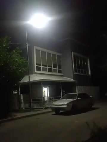 A dark, outdoor scene at night illuminated by a bright streetlamp. A silver sedan is parked on the side of a paved road in front of a two-story building. The building has a dark facade with large, multi-paned windows on the upper floor and a covered porch with railings on the lower floor. A tree with green foliage is visible to the left of the building. The scene is quiet and still.