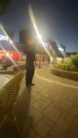 The video shows a person doing a handstand on a brick-paved area outdoors, likely a patio or public space. The person is wearing dark-colored pants and a dark shirt. They execute the handstand, hold it briefly, and then come down. Behind the person are commercial buildings, some of which have visible signage (including "Sotheby's"), indicating a retail or business district. There is also a small garden area with flowers visible. The time appears to be dusk or evening, as the sky is dark but there are streetlights illuminating the area. A portion of a chair is visible in the foreground.