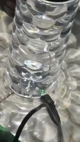 The video shows the base of a cylindrical glass lamp. The lamp is constructed from clear, rounded glass elements that are stacked to form its body. A black USB cable is connected to the base of the lamp. The lamp rests on a white, slightly reflective surface which creates soft, flower-like light patterns around it. The camera is positioned close to the lamp, providing a detailed view of its construction and the cable connection.