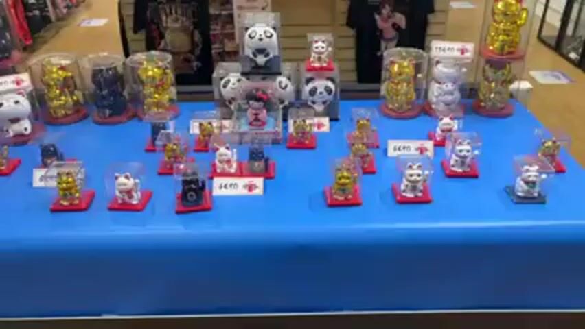 The video shows a table display of various figurines, predominantly of cats, in what appears to be a store setting. The figurines are arranged on a blue tablecloth. Several of the figurines are in clear plastic cases, some elevated on red platforms. A few larger figurines, including panda designs, are also present. Price tags are visible near some items, indicating they are for sale. T-shirts are visible in the background on a display rack. The scene seems to be within a retail environment.