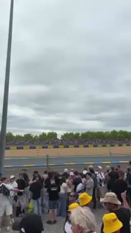 Here's a detailed description of the video:

**Scene:** The video captures a crowd of people gathered alongside a racetrack, likely to watch a race or event. 

**Environment:** The sky is overcast with a grey cloud cover. In the background is the racetrack with a blue and yellow barrier, a grandstand filled with spectators, and a tent. 

**People:** A large crowd is visible in the foreground. They appear to be of mixed ages and are dressed in casual attire. Several people have their backs to the camera, while others are facing the track. There are people wearing hats, some are holding phones, and there's a child riding on a person's shoulders.

**Objects:** In addition to the racetrack and the crowd, a race car speeds past. It is green and moves from left to right in the frame.

**Actions:** The race car zips past the crowd. People are looking towards the track, presumably watching the race. Some people are taking photos or videos with their phones.

**Interactions:** People are standing close to each other, and some appear to be talking to their companions.

**Sounds:** The dominant sound appears to be the roar of the passing race car.
