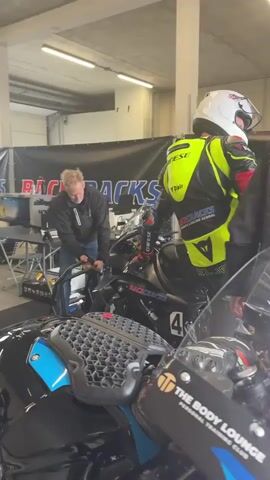 A man wearing a white helmet and a bright yellow and red racing suit is standing next to a motorcycle. Another man in a black jacket is leaning over the motorcycle, appearing to be working on it. In the background, a banner with the words "RACE TRACK" is visible. The scene appears to be a pit area or garage. The racer turns his head and looks towards another person who is partially visible on the right side of the frame.