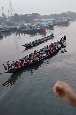 Here is a description of the video:

The video captures a wide shot of a river, possibly taken from a bridge or high vantage point.  The overall scene is a bit hazy or misty, with subdued lighting. Several boats are visible on the river. The main focus is a long, narrow boat crammed with many people, moving across the water. Another similar boat is travelling parallel and slightly behind it.  Other, larger vessels are moored at the edge of the water.

The primary interaction observed is the movement of the crowded boats across the river. The environment includes the river, boats, and a hazy cityscape in the background, with glimpses of buildings, vegetation, and utility poles or towers.