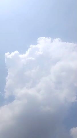 The video shows a vertical shot of a large white cloud against a clear blue sky. The sun appears to be to the left, casting a bright light on the cloud, making the left side very bright and the right side a bit darker and more shadowed. The cloud is the dominant feature, filling most of the frame. The camera seems to be stationary, capturing a still image of the sky and cloud. There are no people or objects visible in the frame, only the natural elements of the sky.