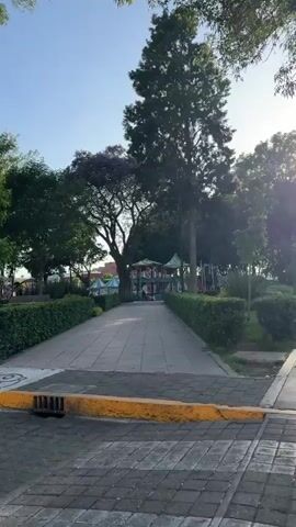 The video captures a series of street scenes alongside a park or public square, likely in Cholula, Mexico. The time of day appears to be sunny, with clear blue skies.

The first shot pans across a cobblestone road with a yellow and blue painted curb, revealing a drain cover. Beyond the curb are neatly trimmed hedges, a paved pathway, and numerous green trees. In the background, colorful playground equipment or small carnival rides are visible.

The scene then shifts to a black motorcycle parked by the curb, with its license plate "72TMF7" partially visible. The park's greenery and structures remain in the background.

Next, the camera is inside a vehicle, showing a red hatchback (a Seat Leon) parked or moving slowly. Its license plate "UAG-81-29" is visible. Behind the car stands a large tree and a plain, rectangular, light-colored monument or base.

The final moments show a white pickup truck, possibly a Chevrolet Silverado, also by the curb, with a colorful ride featuring a character in the background, suggesting a children's play area or a small fair within the park. A sign reading "ZONA DE PARQUIMETRO" (Parking Zone) is also briefly visible.