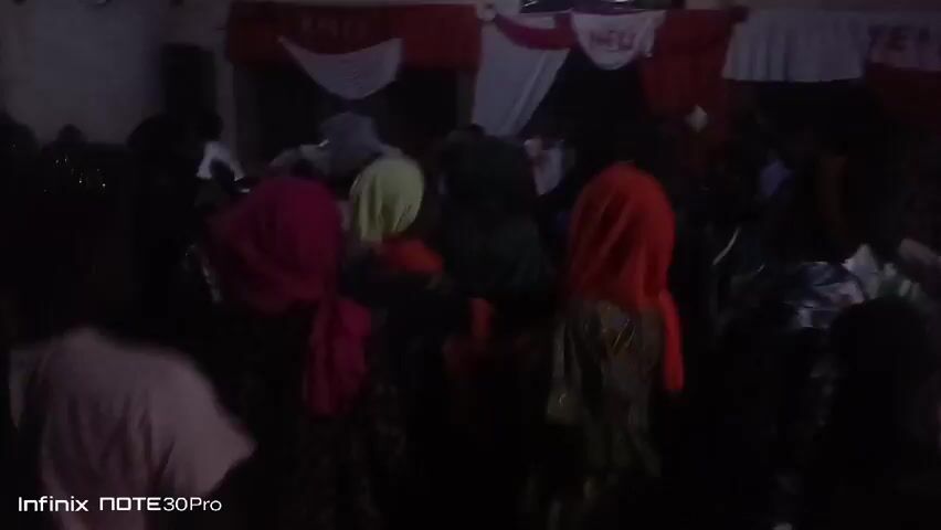 The video shows a group of people gathered in a room, possibly a community hall or event space, likely for a celebration. The lighting is dim. The people are dressed in a variety of colorful clothes and head coverings. There are decorations on the wall behind the group, including red and white drapery. The decorations spell out "NEW YEAR." A speaker is visible in the background. The camera remains mostly stationary, focusing on the crowd.