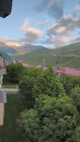 The video shows a wide view of a village nestled in a valley with green hills and mountains in the background. The sky is blue with scattered clouds, some of which are illuminated by the setting sun. In the foreground, there are trees and some buildings with red roofs. A bird flies across the frame from left to right. The overall impression is a serene, rural landscape at dusk.