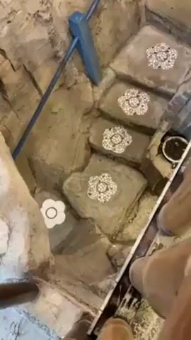 Here is a description of the video:

The video begins with a close-up of an intricately designed staircase. Each step is made of what appears to be sculpted stone with embedded flower-like designs. A blue railing runs alongside the stairs. The camera then pans upwards, revealing the top of the staircase, where there's a mural on the wall depicting a landscape with mountains, water, and greenery. In the background, a doorway leads to a room with chairs. The camera continues to pan up to show a decorative chandelier hanging from the white ceiling. The ceiling has visible support beams.