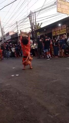 A performer dressed in a red and orange costume with black face paint is performing acrobatic moves in a street. The performer is barefoot and is surrounded by a crowd of onlookers. The scene appears to be in a marketplace or festival setting, with stalls and banners visible in the background. The performer is captured mid-movement, demonstrating a handstand and a rapid spin. The lighting suggests it is daytime.