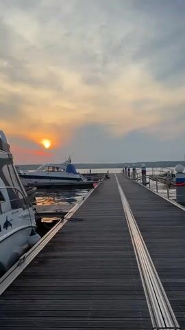 The video pans across a marina at sunset. In the foreground, a wooden pier stretches into the distance, with boats docked on either side. The setting sun casts a warm glow on the water and the sky. The video focuses on the pier and the boats, with a few shots of the water and the surrounding landscape. There are no people visible in the video. The mood is peaceful and serene.