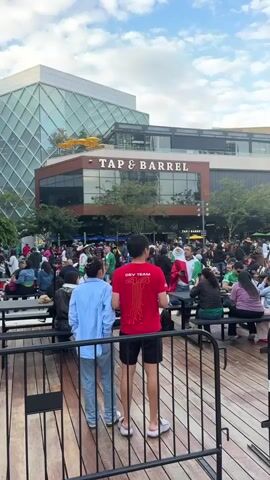 The video shows an outdoor gathering of people in front of a building with "TAP & BARREL" signage. Many people are wearing green and white, suggesting a celebration or event related to Mexico. There is a large crowd, with some individuals seated at tables and others standing. A person in the foreground, wearing a red shirt with "DEV TEAM" on it, is facing away from the camera. In the background, a person wearing a large sombrero and dressed in green and white is visible, holding a white cloth. The atmosphere appears festive, with a cloudy sky overhead.