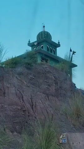 The video shows a green building with a dome situated on a rocky, overgrown hillside under an overcast sky. Flags are visible on the building's roof. Several people are present on or near the building, engaged in what appears to be construction or maintenance work, as evidenced by the presence of tarps, tools, and materials. The surrounding environment is characterized by rough terrain, sparse vegetation, and a slightly hazy atmosphere. The camera appears to be in motion, likely from a vehicle, as it pans across the scene.