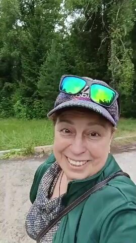 A woman wearing a baseball cap with sunglasses perched on top and a green jacket is smiling at the camera. She is outdoors with trees and tall grass in the background.