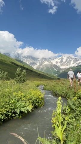 Two people walk away from the camera along a path next to a stream. The path is bordered by green grass and plants. In the background, a majestic snow-capped mountain range is visible under a bright blue sky with fluffy white clouds.
