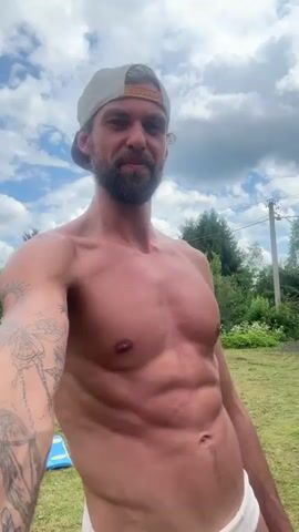 The media file is a selfie video of a shirtless man standing outdoors. He is wearing a beige baseball cap and has a beard. His left arm, visible in the foreground, is adorned with tattoos, including a butterfly design. The man appears to be in good physical shape, with visible abdominal muscles.

The setting is a grassy area with trees and vegetation in the background. There is a blue object, possibly a pool float, on the ground to the left. A utility pole and some fencing are also visible in the distance, suggesting a rural or suburban location. The sky is partly cloudy, indicating daytime, likely late morning or afternoon, with fair weather. The man is looking directly at the camera with a relaxed expression. There is no visible text in the media file.