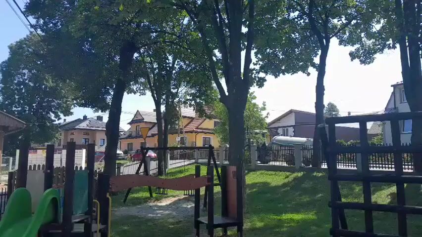 The video shows a playground area with trees and houses in the background. In the foreground, there are several pieces of playground equipment, including a green slide, swings, and a climbing structure. The ground is covered in sand and grass. The houses in the background appear to be residential. The sky is visible and appears to be blue and clear. The scene is well-lit, suggesting it is daytime.