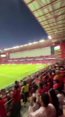 The video depicts a crowded stadium at night. A large number of people fill the stands, mostly wearing red clothing. The camera angle is from within the crowd, providing a close view of people cheering and raising their hands. A soccer field is visible below, illuminated by bright lights, with a game in progress. The stadium has a red roof structure and the sky is a dark shade of blue. The overall atmosphere suggests a lively and energetic event.