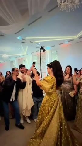 A group of people are gathered in a banquet hall, with one woman in a golden dress dancing in the center. She is wearing a crown and appears to be smiling and enjoying herself. Several people are filming her with their phones. The hall is decorated with white drapes and chandeliers. The lighting is bright and festive. The woman's dress has puffed sleeves and a long train. The people around her are clapping and cheering. The man in the front left is wearing a suit and jeans and is also clapping.