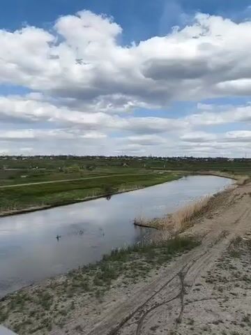 The video presents a panoramic view of a rural landscape under a partly cloudy sky. A narrow river or canal flows from the left side of the frame towards the right, reflecting the bright sky. Reeds and other vegetation line its banks. To the immediate right of the river, there's a dirt or sandy track. Further right, a vast, freshly tilled field of dark brown earth stretches into the distance, showing distinct furrows. Beyond the river and the tilled field, green fields and grasslands extend towards a distant horizon. A village or small town, characterized by various buildings and what appear to be church steeples or towers, is visible on the horizon line in the center and right. The sky is a vibrant blue, adorned with numerous fluffy white cumulus clouds, suggesting a pleasant daytime setting. There are no people visible, and no discernible text appears in the frame. The overall scene depicts a calm, agricultural area, consistent with the Inhulets region of Ukraine.
