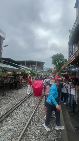 The video shows a crowded street lined with shops and market stalls, with people walking along a railway track that runs down the middle of the street. A red sky lantern is being held up by two people, and another sky lantern is already floating in the air above them. The sky is overcast.