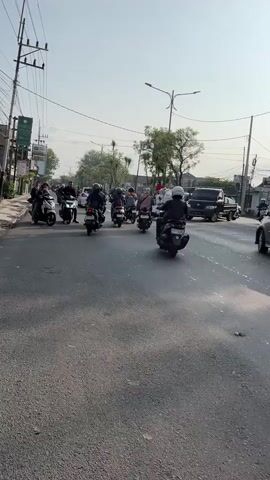 The media file depicts a street scene with several people riding motorcycles. The subjects are a mix of individuals, some appearing to be riding alone, while others are in groups on the same motorcycles. The objects visible include numerous motorcycles of various models, a black pickup truck in the background, and utility poles with wires. The scene is set on a paved road with sidewalks and buildings lining one side. Trees are also present, suggesting an outdoor environment.

The activity taking place is traffic movement, with the motorcycles traveling along the road. Some are moving at a steady pace, while others seem to be in a cluster, possibly at an intersection or a point of congestion.

Notable details include the time of day, which appears to be morning or early afternoon, given the bright daylight and the shadows cast. The weather seems clear and sunny, with a bright sky. Location cues are subtle but suggest an urban or semi-urban setting, possibly in Southeast Asia, given the style of motorcycles and buildings. There are no strong indicators of specific emotions, as the focus is on the activity of riding.

Visible text includes signage on the buildings and poles, but it is largely illegible due to the distance and resolution of the media file. One green sign on a pole has some text on it, but it is too blurry to read. The overall impression is of a typical day with motorcycle traffic in a populated area.