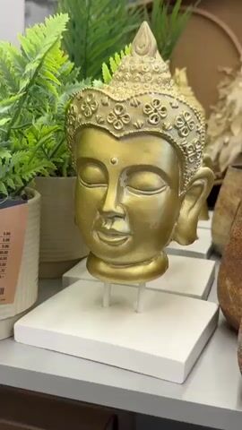 The video shows a close-up of a golden Buddha head statue. The statue is placed on a white stand. In the background, there are green plants and other decorative items. The camera slowly zooms in on the statue's face, highlighting the intricate details of its headdress and facial features. The lighting is soft and warm, creating a serene and tranquil atmosphere. The video appears to be filmed in a store or a home decor setting.
