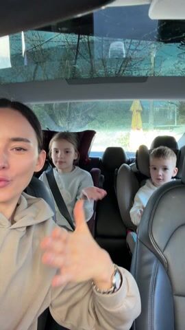 A woman records a video from the driver’s seat of a car. Two children are in the back seat. A girl sits behind the driver, and a toddler is in a child safety seat behind the passenger seat. The woman wears a beige hoodie and has dark hair pulled back. She gestures and appears to be singing along to music playing in the car. The children appear to be clapping along. A glass roof is visible above the passengers in the car, through which a tree and a yellow umbrella can be seen. The interior of the car is black and grey.