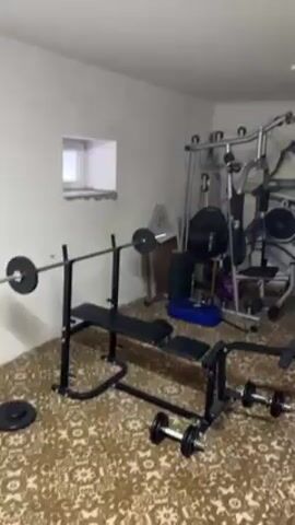 The video shows a home gym room. It has a weight bench, a multi-gym machine, dumbbells, and an airplane mural on the wall. There is also a flag with the colors red, yellow, and black. A piece of paper with handwritten text in Russian is pinned to a corkboard. The camera pans around the room, showing the equipment and details.