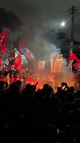 The video shows a nighttime protest taking place outdoors. A large crowd of people are gathered around a fire.  They are holding flags and banners, some of which are red and appear to be associated with UNESP. There is a lot of smoke from the fire. The crowd is dense, with many faces visible as people move around and raise their arms. The scene is lit by streetlights and the fire.  Some people are taking photos or videos of the event with their phones.  The atmosphere appears to be intense and energetic.