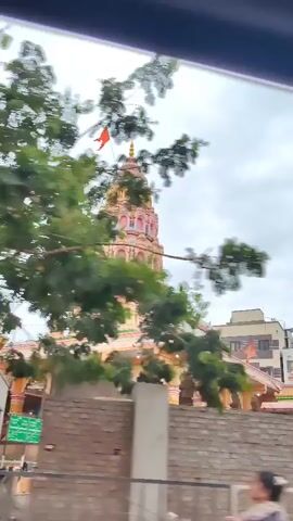 A video shows a Hindu temple on a cloudy day. The temple has a tall, colorful spire topped with a flag. There are arched walkways and decorative elements on the temple's exterior. In the foreground, a brick wall and some vegetation are visible. The overall impression is of a religious structure viewed from a distance, possibly from a moving vehicle.