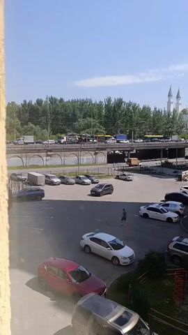 The video shows a parking lot with several cars. A man is walking in the parking lot. There is a road in the background with traffic. There are also buildings and trees in the background. The sky is blue with some clouds.