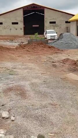 The video pans across a construction site. In the background, a large warehouse-like building with an open entrance stands. To the right of the warehouse is a smaller building with a yellow roof and multiple windows. A white pickup truck is parked in front of the warehouse. A large pile of gravel is next to the pickup truck. The foreground shows a dirt and gravel area with piles of dirt, some scattered bricks, and a yellow hose running across the ground. There is evidence of recent rain with puddles of water visible. No people are clearly visible in the video. The scene appears to be a work in progress with construction materials and equipment present.